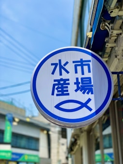 A round sign with blue Japanese characters and a stylized fish symbol against a white background is displayed outside. It is mounted on the side of a building, with an urban street scene partially visible in the background and a clear blue sky above.