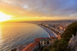 A coastal cityscape at sunset with vibrant orange and yellow hues illuminating the sky. The view captures a sweeping stretch of coastline with buildings lining the shore, and the sun setting over the calm sea. Hills are visible in the background, creating a serene and picturesque atmosphere.