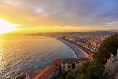 A coastal cityscape at sunset with vibrant orange and yellow hues illuminating the sky. The view captures a sweeping stretch of coastline with buildings lining the shore, and the sun setting over the calm sea. Hills are visible in the background, creating a serene and picturesque atmosphere.