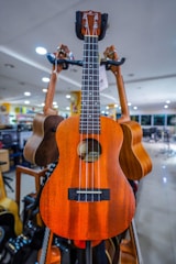 A wooden ukulele with four strings is prominently displayed in a well-lit music store. The background includes other guitars hanging and bright overhead lights.