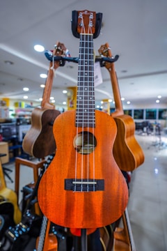 A wooden ukulele with four strings is prominently displayed in a well-lit music store. The background includes other guitars hanging and bright overhead lights.