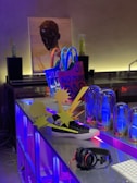 A modern and eclectic setup featuring a brightly colored blue and yellow tote bag with bold text, a black sneaker with white soles, headphones resting on a metallic surface, and decorative glass domes illuminated with blue lighting. A portrait of a head is visible in the background, adding an artistic touch.