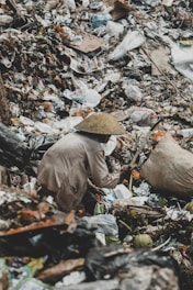 A person wearing a conical hat is foraging through a large pile of garbage. The scene is cluttered with various discarded items, including plastic bags, cardboard, and other debris.