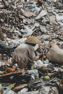 A person wearing a conical hat is foraging through a large pile of garbage. The scene is cluttered with various discarded items, including plastic bags, cardboard, and other debris.