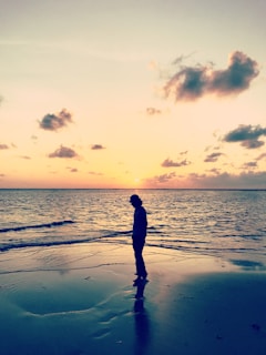 A silhouette of a person standing on a beach at sunset, looking out over calm waves, symbolizing reflection and hope.