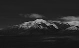 A striking black and white photograph of a mountain range with snow-capped peaks. Shadows and light play across the rugged landscape, creating a dramatic contrast against the dark sky. The clouds appear soft and slightly illuminated, adding texture to the scene.