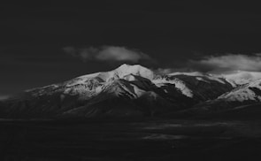 A striking black and white photograph of a mountain range with snow-capped peaks. Shadows and light play across the rugged landscape, creating a dramatic contrast against the dark sky. The clouds appear soft and slightly illuminated, adding texture to the scene.