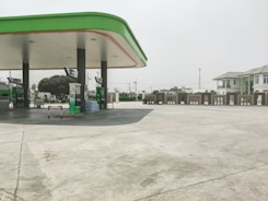 A clean, empty gas station with a green and white color scheme. The station has multiple fuel pumps topped with an extended canopy and is surrounded by a wide concrete area. In the background, there are trees, utility poles, and a multi-story building with a light gray exterior.