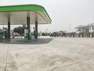 A clean, empty gas station with a green and white color scheme. The station has multiple fuel pumps topped with an extended canopy and is surrounded by a wide concrete area. In the background, there are trees, utility poles, and a multi-story building with a light gray exterior.
