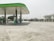 A clean, empty gas station with a green and white color scheme. The station has multiple fuel pumps topped with an extended canopy and is surrounded by a wide concrete area. In the background, there are trees, utility poles, and a multi-story building with a light gray exterior.