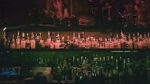 Rows of liquor bottles are lined up on shelves in a dimly lit bar, with a warm, moody ambiance. The shelves are adorned with mixed alcoholic brands, and the decor includes dark wood and artistic portraits, contributing to an intimate atmosphere.
