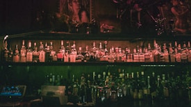 Rows of liquor bottles are lined up on shelves in a dimly lit bar, with a warm, moody ambiance. The shelves are adorned with mixed alcoholic brands, and the decor includes dark wood and artistic portraits, contributing to an intimate atmosphere.