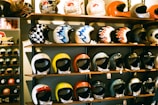 A display of colorful motorcycle helmets arranged on wooden shelves, showcasing various designs and styles. The helmets feature artistic patterns, including checkerboard and flame motifs, and are placed on multiple levels.