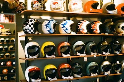 A display of colorful motorcycle helmets arranged on wooden shelves, showcasing various designs and styles. The helmets feature artistic patterns, including checkerboard and flame motifs, and are placed on multiple levels.