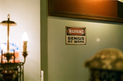 A dimly lit room featuring a vintage decor lamp and a wall sign that reads 'WARNING GENIUS AT WORK'. The background includes a softly illuminated wall with some wooden paneling.