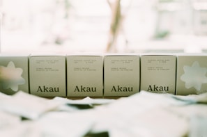 Five boxes with the brand name Akau displayed on a surface, each indicating they contain steeped coffee bags with specifications of weight and pack size. The background appears bright and out of focus, creating a soft, muted tone.