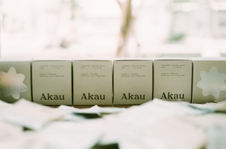 Five boxes with the brand name Akau displayed on a surface, each indicating they contain steeped coffee bags with specifications of weight and pack size. The background appears bright and out of focus, creating a soft, muted tone.