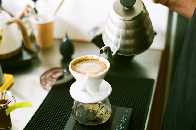 A barista is pouring hot water from a stainless steel gooseneck kettle into a white ceramic dripper containing coffee grounds. The setup includes a scale beneath the dripper and a glass carafe collecting the brewed coffee. Various other coffee-making tools and a cup are visible in the background.