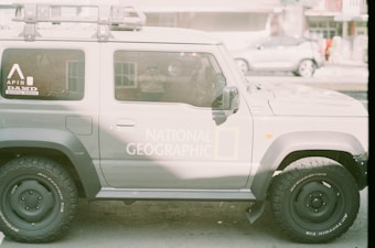 A grey SUV with the National Geographic logo on the side is parked on the street. The vehicle features accessories such as a roof rack and rugged tires. The background includes blurred images of other vehicles and a building, giving an urban setting.