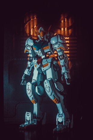 A detailed and intricately designed robot or mecha stands in a dramatic pose, highlighted by a combination of blue, white, and orange armor plating. The background is dark, with subtle geometric patterns illuminated in warm tones, giving a futuristic and sci-fi ambiance.