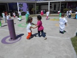 Children are playing outdoors on a sunny day, engaging in various activities like running and interacting in groups. The area is colorful with various play structures and painted circles on the ground, suggesting it is a playground or schoolyard.