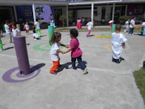 Children are playing outdoors on a sunny day, engaging in various activities like running and interacting in groups. The area is colorful with various play structures and painted circles on the ground, suggesting it is a playground or schoolyard.