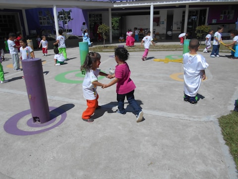Children are playing outdoors on a sunny day, engaging in various activities like running and interacting in groups. The area is colorful with various play structures and painted circles on the ground, suggesting it is a playground or schoolyard.