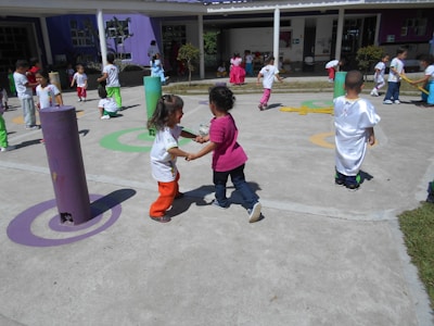 Children are playing outdoors on a sunny day, engaging in various activities like running and interacting in groups. The area is colorful with various play structures and painted circles on the ground, suggesting it is a playground or schoolyard.