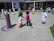 Children are playing outdoors on a sunny day, engaging in various activities like running and interacting in groups. The area is colorful with various play structures and painted circles on the ground, suggesting it is a playground or schoolyard.