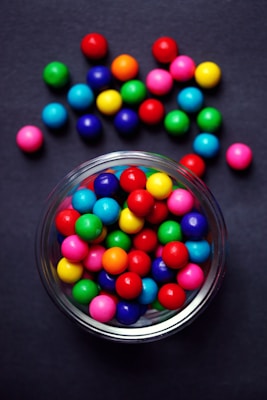 A clear glass bowl filled with bright, multicolored gumballs is placed against a dark background. The gumballs display a variety of colors including red, blue, green, yellow, orange, and pink. Some gumballs are scattered outside the bowl, enhancing the vibrant and playful aesthetic.