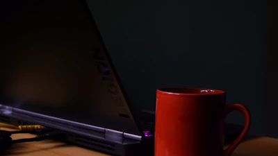 A cozy corner with a black and red mug steaming beside an open laptop displaying the blog homepage.