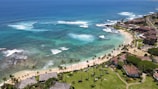 A picturesque coastal scene featuring turquoise ocean waters with gentle waves breaking near the shore. The coastline is lined with sandy beaches and bordered by lush greenery and palm trees. Residential buildings and resorts are visible along the coast, with roads and pathways leading through the area.