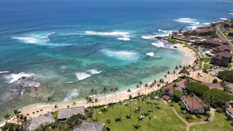 A picturesque coastal scene featuring turquoise ocean waters with gentle waves breaking near the shore. The coastline is lined with sandy beaches and bordered by lush greenery and palm trees. Residential buildings and resorts are visible along the coast, with roads and pathways leading through the area.