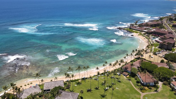 A picturesque coastal scene featuring turquoise ocean waters with gentle waves breaking near the shore. The coastline is lined with sandy beaches and bordered by lush greenery and palm trees. Residential buildings and resorts are visible along the coast, with roads and pathways leading through the area.