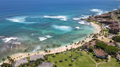 A picturesque coastal scene featuring turquoise ocean waters with gentle waves breaking near the shore. The coastline is lined with sandy beaches and bordered by lush greenery and palm trees. Residential buildings and resorts are visible along the coast, with roads and pathways leading through the area.