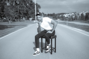 man in white t-shirt and black pants sitting on black wooden chair on road during