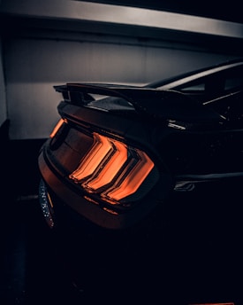 A rear view of a car's taillights illuminated with a vibrant orange glow, set against a dark, shadowy background. The taillights feature a distinctive chevron pattern, highlighting the sleek and modern design of the vehicle.