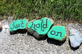 Three stones are painted green with the words 'Just hold on' written in black. They are placed on a gravel path next to a patch of grass.