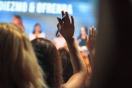 A raised hand holding a pen, with a blurred background of people who appear to be in an audience or gathering. The focus is on the hand in the foreground, while people in the background are slightly out of focus, suggesting a setting like a conference or meeting.