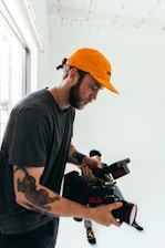 A man with tattoos is holding a professional video camera. He wears an orange cap and a dark t-shirt. Another person, slightly out of focus, sits on the floor in the background against a white wall.