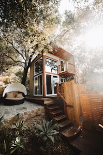 A two-story wooden tiny house nestled among trees, with large windows and a small balcony. The structure features a set of wooden stairs leading up to it. Nearby, there is a cozy outdoor seating area with a round, white canopy and cushions. The surrounding area includes plants and a stone-paved path, giving a rustic and natural atmosphere.