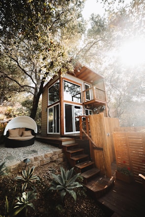 A two-story wooden tiny house nestled among trees, with large windows and a small balcony. The structure features a set of wooden stairs leading up to it. Nearby, there is a cozy outdoor seating area with a round, white canopy and cushions. The surrounding area includes plants and a stone-paved path, giving a rustic and natural atmosphere.