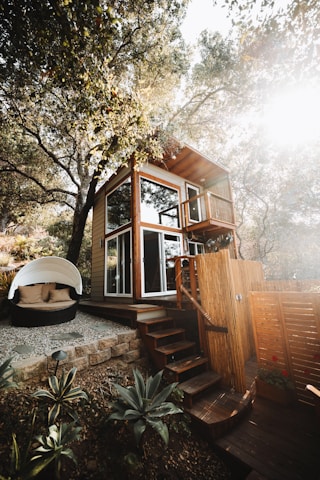 A two-story wooden tiny house nestled among trees, with large windows and a small balcony. The structure features a set of wooden stairs leading up to it. Nearby, there is a cozy outdoor seating area with a round, white canopy and cushions. The surrounding area includes plants and a stone-paved path, giving a rustic and natural atmosphere.