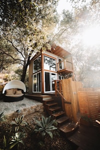 A two-story wooden tiny house nestled among trees, with large windows and a small balcony. The structure features a set of wooden stairs leading up to it. Nearby, there is a cozy outdoor seating area with a round, white canopy and cushions. The surrounding area includes plants and a stone-paved path, giving a rustic and natural atmosphere.