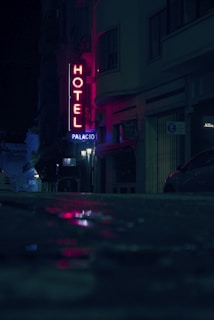 A dimly lit urban street at night featuring a neon hotel sign with the word 'Hotel' in red and 'Palacio' beneath it. The street is wet, reflecting the neon lights, creating a mysterious and moody atmosphere. Buildings and an indistinct figure are visible in the background along with parked cars.