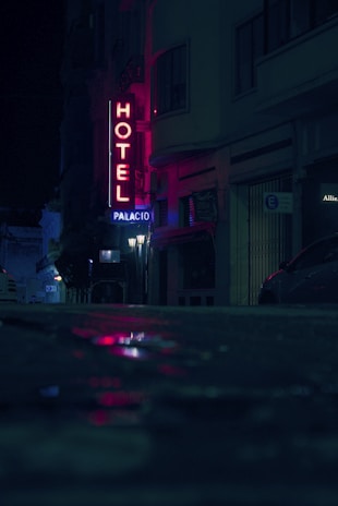 A dimly lit urban street at night featuring a neon hotel sign with the word 'Hotel' in red and 'Palacio' beneath it. The street is wet, reflecting the neon lights, creating a mysterious and moody atmosphere. Buildings and an indistinct figure are visible in the background along with parked cars.