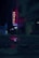 A dimly lit urban street at night featuring a neon hotel sign with the word 'Hotel' in red and 'Palacio' beneath it. The street is wet, reflecting the neon lights, creating a mysterious and moody atmosphere. Buildings and an indistinct figure are visible in the background along with parked cars.