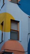 A building exterior with two windows features a vibrant geometric mural. Large, colorful circles in yellow, orange, and blue cover the surface, creating an eye-catching design. The window frames are painted in a light blue shade, and there are visible electrical wires running along the wall.