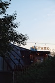 A rooftop venue with a sign in Cyrillic script, partially obscured by foliage. The structure features a sloped roof and string lights hanging overhead. The sky is clear and softly lit during sunset, casting a serene ambiance.