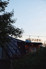 A rooftop venue with a sign in Cyrillic script, partially obscured by foliage. The structure features a sloped roof and string lights hanging overhead. The sky is clear and softly lit during sunset, casting a serene ambiance.